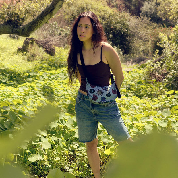 A lifestyle image of a person wearing a small Crescent Bag from Baggu in Digital Denim Birds surrounded by plants