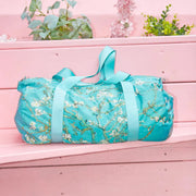 A light blue LOQI weekender bag featuring Van Gogh's Almond Blossom artwork. The bag is shown on a pink bench with flowers behind. 